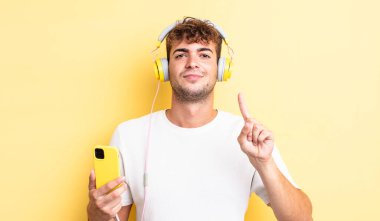 Genç yakışıklı adam gülümsüyor ve arkadaş canlısı görünüyor, bir numarayı gösteriyor. kulaklıklar ve akıllı telefon konsepti