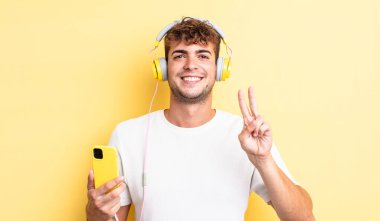 Genç yakışıklı adam gülümsüyor ve arkadaş canlısı görünüyor, iki numarayı gösteriyor. kulaklıklar ve akıllı telefon konsepti