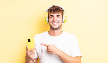 Genç yakışıklı adam neşeyle gülümsüyor, mutlu hissediyor ve bir tarafı işaret ediyor. kulaklıklar ve akıllı telefon konsepti