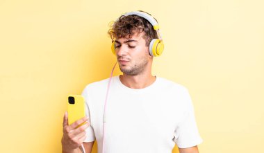 Genç ve yakışıklı bir adam üzgün, üzgün ya da kızgın hissediyor ve yan tarafa bakıyor. kulaklıklar ve akıllı telefon konsepti