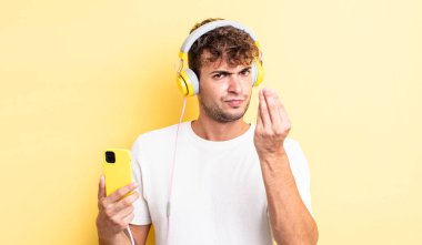 Genç yakışıklı adam kapiş ya da para hareketi yapıyor, sana ödemeni söylüyor. kulaklıklar ve akıllı telefon konsepti