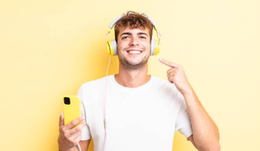 Genç ve yakışıklı bir adam kendi gülüşünü işaret ederek kendine güvenerek gülümsüyor. kulaklıklar ve akıllı telefon konsepti