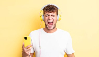 Genç yakışıklı adam agresif bir şekilde bağırıyor, çok kızgın görünüyor. kulaklıklar ve akıllı telefon konsepti