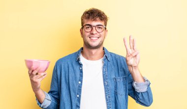 Genç yakışıklı adam gülümsüyor ve arkadaş canlısı görünüyor, üç numarayı gösteriyor. boş kase kavramı