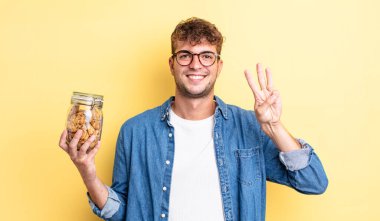 Genç yakışıklı adam gülümsüyor ve arkadaş canlısı görünüyor, üç numarayı gösteriyor. kurabiye şişesi konsepti