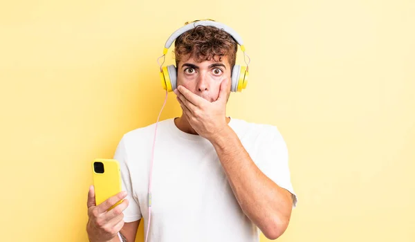 若いハンサムな男がショックで手で口を覆っている ヘッドフォンとスマートフォンのコンセプト — ストック写真