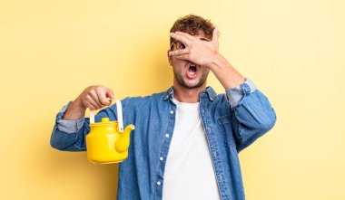 Genç yakışıklı adam şok olmuş, korkmuş ya da korkmuş görünüyor, yüzünü el yapımı çaydanlık konseptiyle kaplıyor.