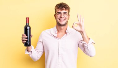Genç yakışıklı adam kendini mutlu hissediyor, iyi bir jestle onaylıyor. şarap şişesi konsepti