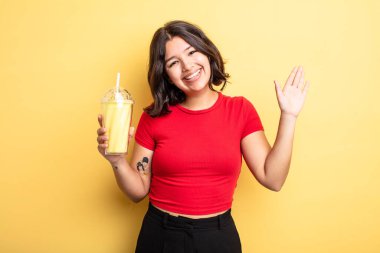 Genç ve güzel bir bayan mutlu bir şekilde gülümsüyor, el sallıyor, sizi selamlıyor ve selamlıyor. Milkshake konsepti