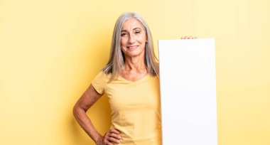 Orta yaşlı güzel bir kadın mutlu bir şekilde gülümsüyor, eli kalçasında ve kendinden emin. boş tuval kavramı