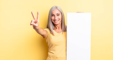 Orta yaşlı güzel bir kadın gülümsüyor ve mutlu görünüyor, zafer ya da barış işareti yapıyor. boş tuval kavramı