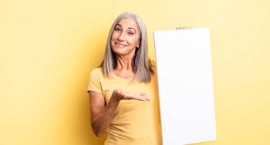 Orta yaşlı güzel bir kadın neşeli bir şekilde gülümsüyor, mutlu hissediyor ve bir konsept gösteriyor. boş tuval kavramı