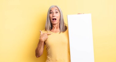 Orta yaşlı güzel bir kadın şok olmuş ve ağzı açık, kendini işaret eden bir şekilde şaşırmış görünüyor. boş tuval kavramı