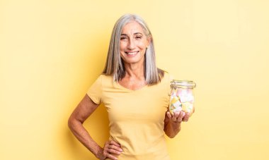 Orta yaşlı güzel bir kadın mutlu bir şekilde gülümsüyor, eli kalçasında ve kendinden emin. Şeker şişesi konsepti