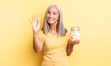 Orta yaşlı, güzel bir kadın mutlu bir şekilde gülümsüyor, el sallıyor, sizi selamlıyor. Şeker şişesi konsepti