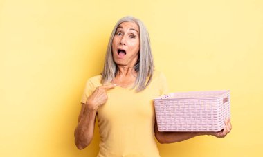 Orta yaşlı güzel bir kadın şok olmuş ve ağzı açık, kendini işaret eden bir şekilde şaşırmış görünüyor. boş sepet konsepti