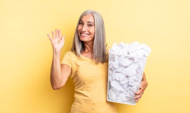 Orta yaşlı, güzel bir kadın mutlu bir şekilde gülümsüyor, el sallıyor, sizi selamlıyor. kağıt toplar başarısızlık konsepti