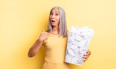 Orta yaşlı güzel bir kadın şok olmuş ve ağzı açık, kendini işaret eden bir şekilde şaşırmış görünüyor. kağıt toplar başarısızlık konsepti