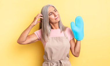 Orta yaşlı, güzel bir kadın şaşkın ve kafası karışık, kaşınıyor. fırın eldiveni kavramı