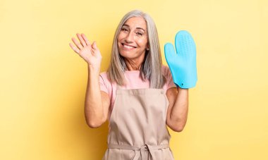 Orta yaşlı, güzel bir kadın mutlu bir şekilde gülümsüyor, el sallıyor, sizi selamlıyor. fırın eldiveni kavramı