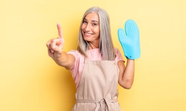 Orta yaşlı güzel bir kadın gülümsüyor ve arkadaş canlısı görünüyor. Bir numarayı gösteriyor. fırın eldiveni kavramı