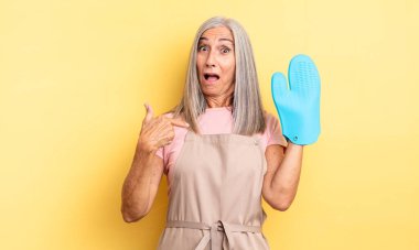 Orta yaşlı güzel bir kadın şok olmuş ve ağzı açık, kendini işaret eden bir şekilde şaşırmış görünüyor. fırın eldiveni kavramı