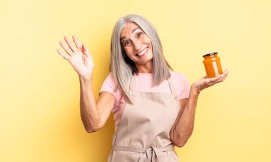 Orta yaşlı, güzel bir kadın mutlu bir şekilde gülümsüyor, el sallıyor, sizi selamlıyor. Şeftali reçeli