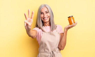 Orta yaşlı güzel bir kadın gülümsüyor ve arkadaş canlısı görünüyor, üç numarayı gösteriyor. Şeftali reçeli