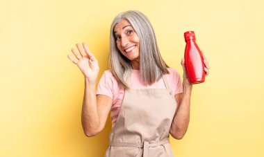 Orta yaşlı, güzel bir kadın mutlu bir şekilde gülümsüyor, el sallıyor, sizi selamlıyor. Ketçap konsepti