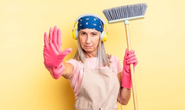 Orta yaşlı, güzel bir kadın ciddi görünüyor. Avuç içini gösteriyor. Durdurma hareketi yapıyor. ev ve süpürge kavramı