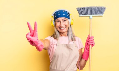 Orta yaşlı güzel bir kadın gülümsüyor ve mutlu görünüyor, zafer ya da barış işareti yapıyor. ev ve süpürge kavramı