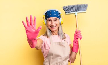 Orta yaşlı güzel bir kadın gülümsüyor ve arkadaş canlısı görünüyor, beş numarayı gösteriyor. ev ve süpürge kavramı