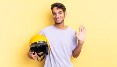 İspanyol yakışıklı adam mutlu bir şekilde gülümsüyor, el sallıyor, seni selamlıyor ve selamlıyor. Motosiklet kaskı konsepti