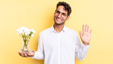 İspanyol yakışıklı adam mutlu bir şekilde gülümsüyor, el sallıyor, seni selamlıyor ve selamlıyor. çiçek saksı konsepti