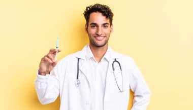 İspanyol yakışıklı adam mutlu bir şekilde gülümsüyor, eli kalçasında ve kendine güvenen bir doktor ve srynge konseptiyle.