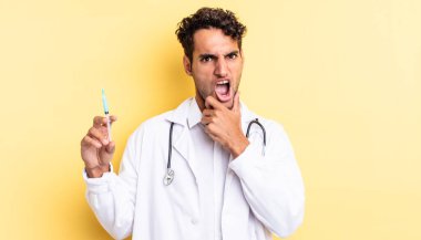 Ağzı ve gözleri sonuna kadar açık ve eli çene hekimi ve srynge konseptinde olan İspanyol asıllı yakışıklı adam.