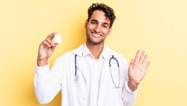 İspanyol yakışıklı adam mutlu bir şekilde gülümsüyor, el sallıyor, seni selamlıyor ve selamlıyor. Hekim şişesi hapları konsepti