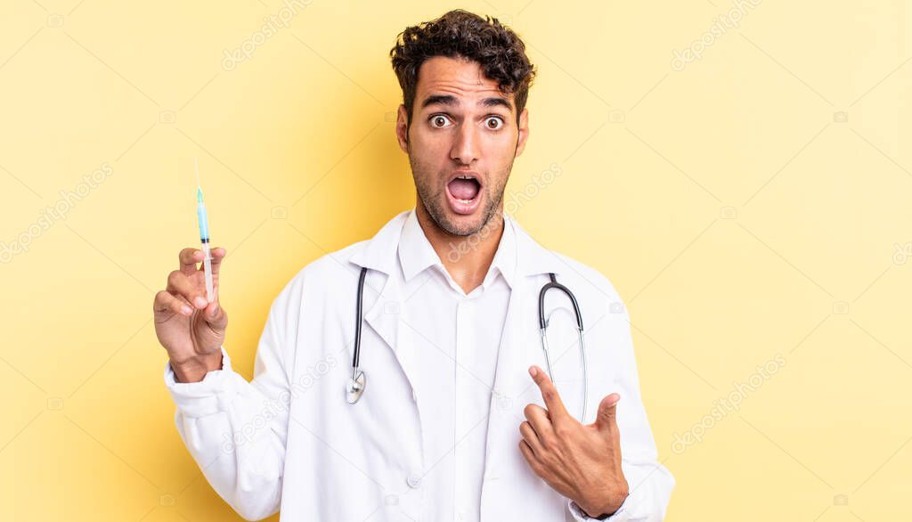 hispanic handsome man looking shocked and surprised with mouth wide open, pointing to self physician and srynge concept