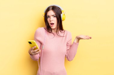 Genç İspanyol kadın şaşkın, şaşkın ve şüpheci hissediyor. kulaklıklar ve telefon konsepti