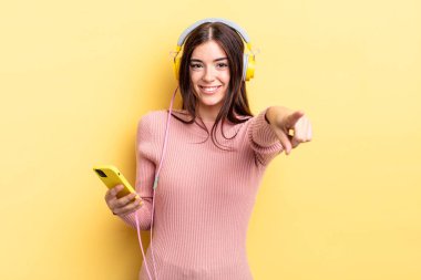 Genç İspanyol kadın kamerayı göstererek seni seçiyor. kulaklıklar ve telefon konsepti