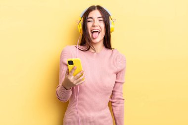 Neşeli ve asi tavırları olan, şaka yapan ve dilini dışarı çıkaran genç İspanyol bir kadın. kulaklıklar ve telefon konsepti