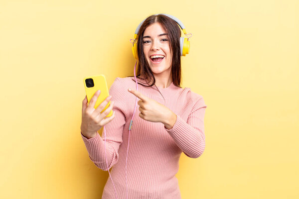 young hispanic woman looking excited and surprised pointing to the side. headphones and telephone concept
