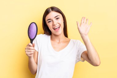 Genç İspanyol kadın mutlu bir şekilde gülümsüyor, el sallıyor, sizi selamlıyor ve selamlıyor. saç fırçası konsepti