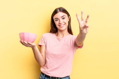 Genç İspanyol kadın gülümsüyor ve arkadaş canlısı görünüyor, üç numarayı gösteriyor. boş kase kavramı