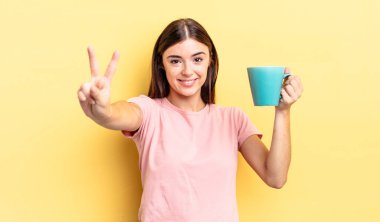 Genç İspanyol kadın gülümsüyor ve arkadaş canlısı görünüyor, iki numarayı gösteriyor. Kahve fincanı konsepti