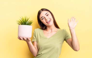 Genç İspanyol kadın mutlu bir şekilde gülümsüyor, el sallıyor, sizi selamlıyor ve selamlıyor. bitki otu konsepti