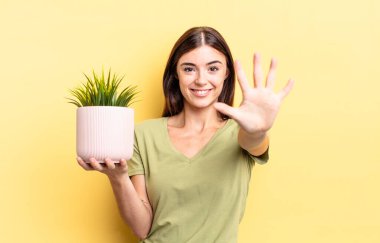 Genç İspanyol kadın gülümsüyor ve arkadaş canlısı görünüyor, beş numarayı gösteriyor. bitki otu konsepti