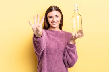 Genç İspanyol kadın gülümsüyor ve arkadaş canlısı görünüyor, dört numarayı gösteriyor. su şişesi kavramı