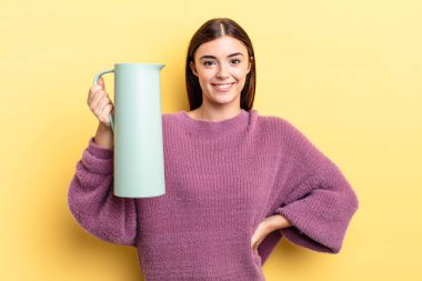 Genç İspanyol kadın mutlu bir şekilde gülümsüyor, eli kalçasında ve kendinden emin. Kahve termosu kavramı