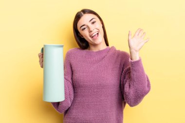Genç İspanyol kadın mutlu bir şekilde gülümsüyor, el sallıyor, sizi selamlıyor ve selamlıyor. Kahve termosu kavramı